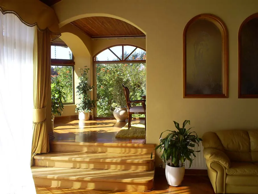 Interior view of a sunlit living room with wooden floors, steps leading to a sunroom with garden view, and a yellow sofa.