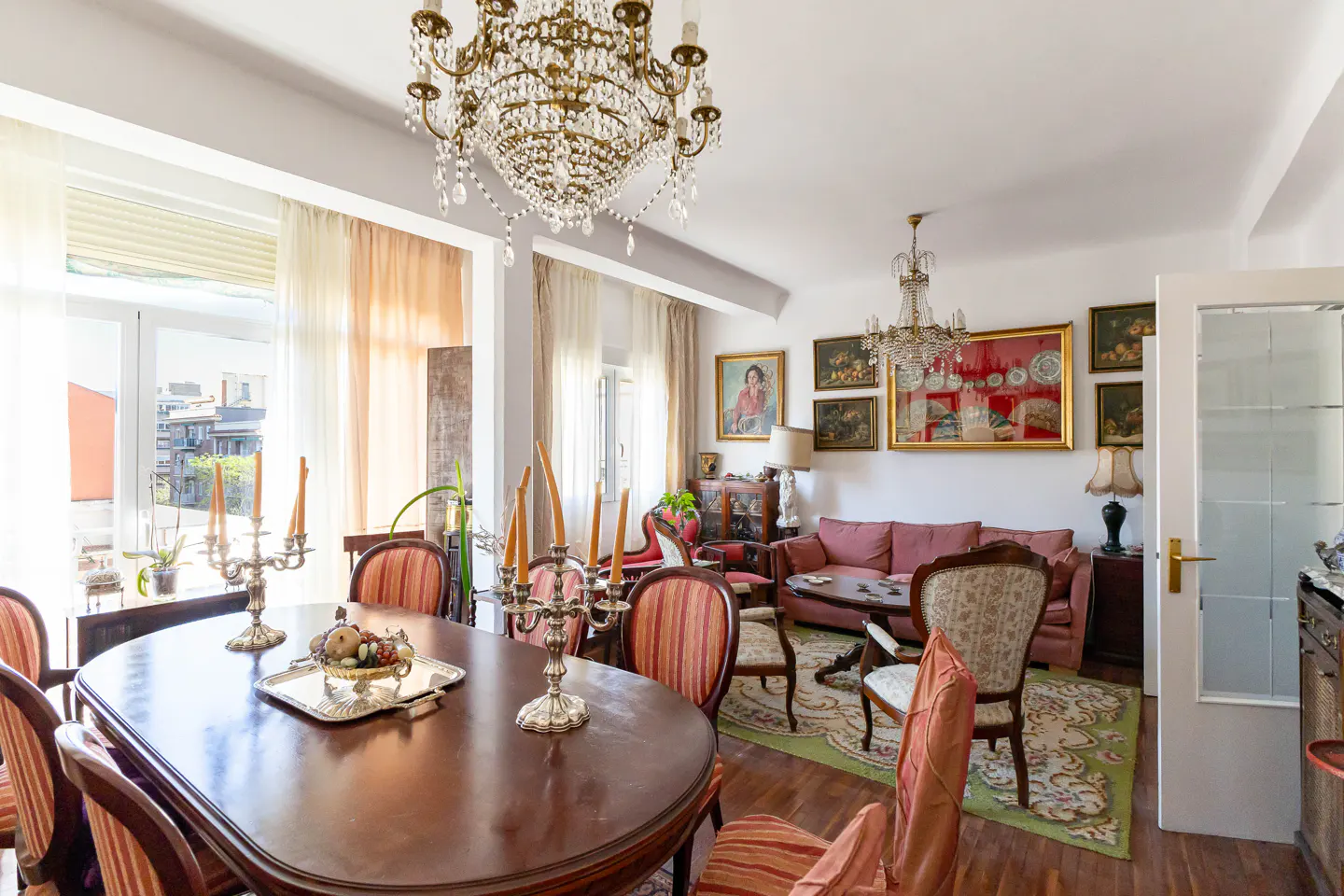 A bright living and dining room with a large wooden table, chairs, and a crystal chandelier. Paintings adorn the walls, and a pink sofa sits in the background.