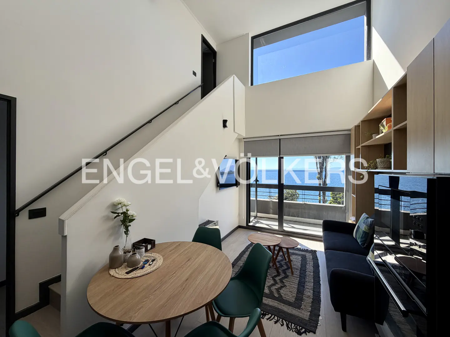 Bright, modern apartment interior with a round wood table, green chairs, and a black sofa facing a balcony with ocean views. Stairs lead to an upper level.