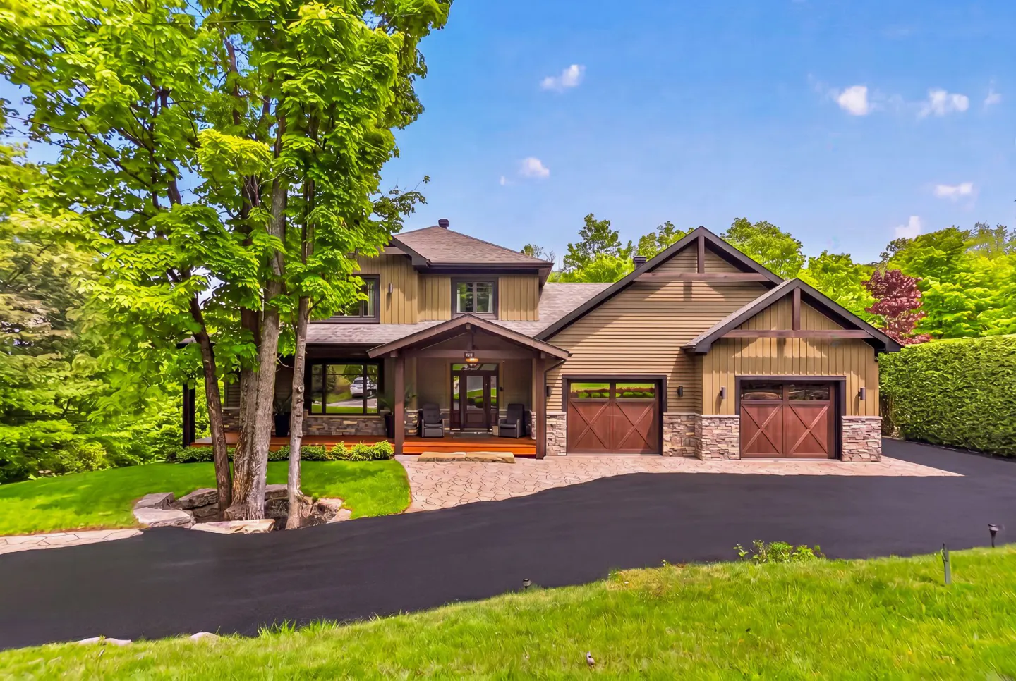Two-story tan home with stone accents, a covered porch, and a two-car garage, set on a lush green lot with a paved driveway.