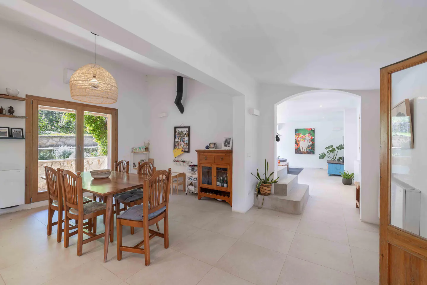 Bright dining room with a wooden table, six chairs, and a straw pendant light. A window overlooks a green garden. The room leads to a living area.