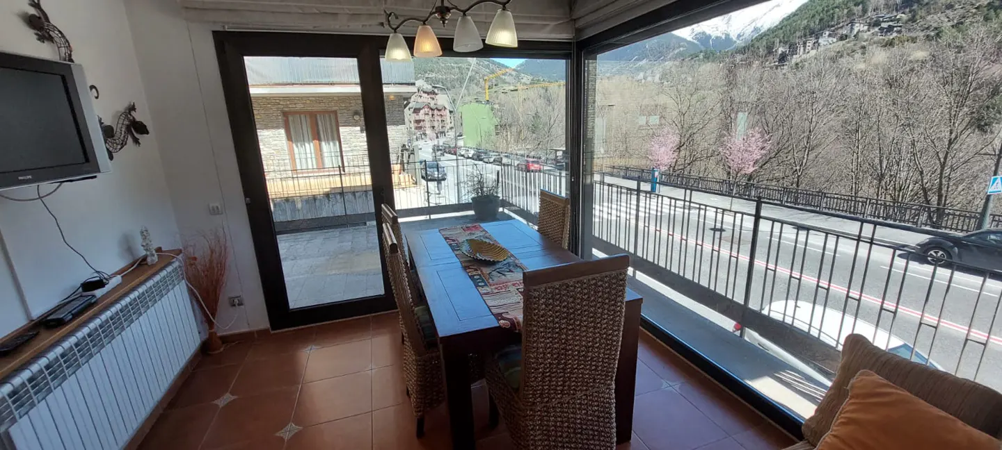 Dining area with a wooden table and wicker chairs, overlooking a street with mountains in the background.