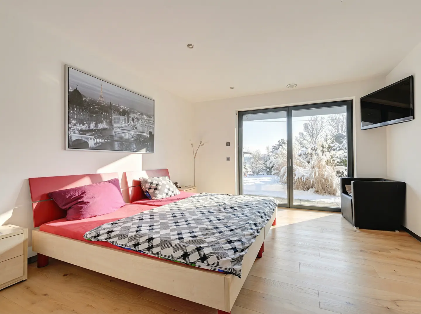 Modern bedroom with a red-framed bed, checkered bedding, Paris wall art, and sliding doors overlooking a snowy garden.