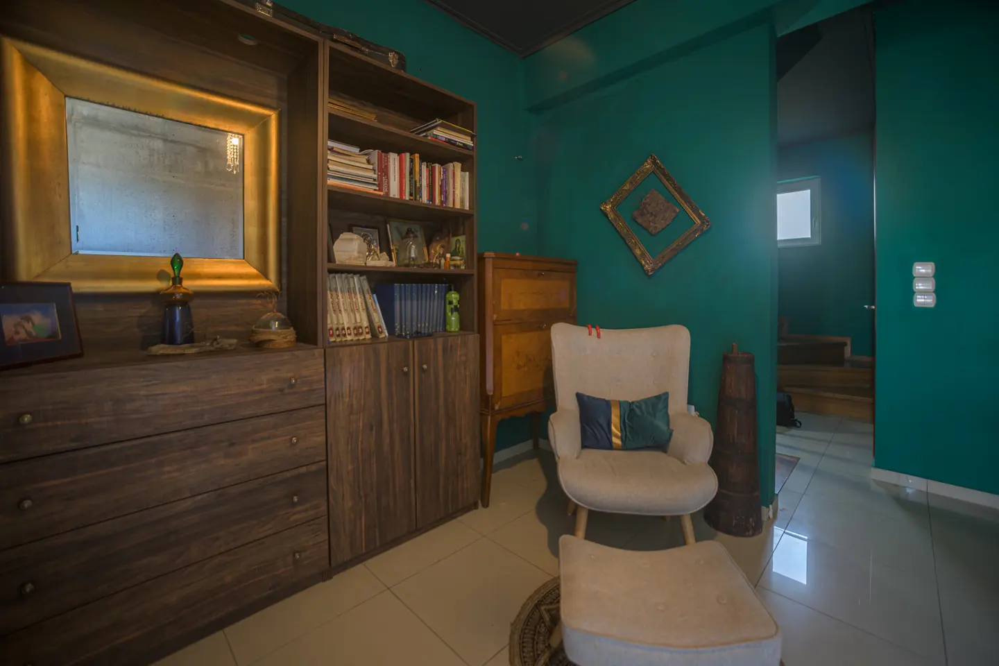 Cozy reading nook with a cream chair, footstool, and dark wood bookshelf against teal walls. A gold-framed mirror and doorway add depth.