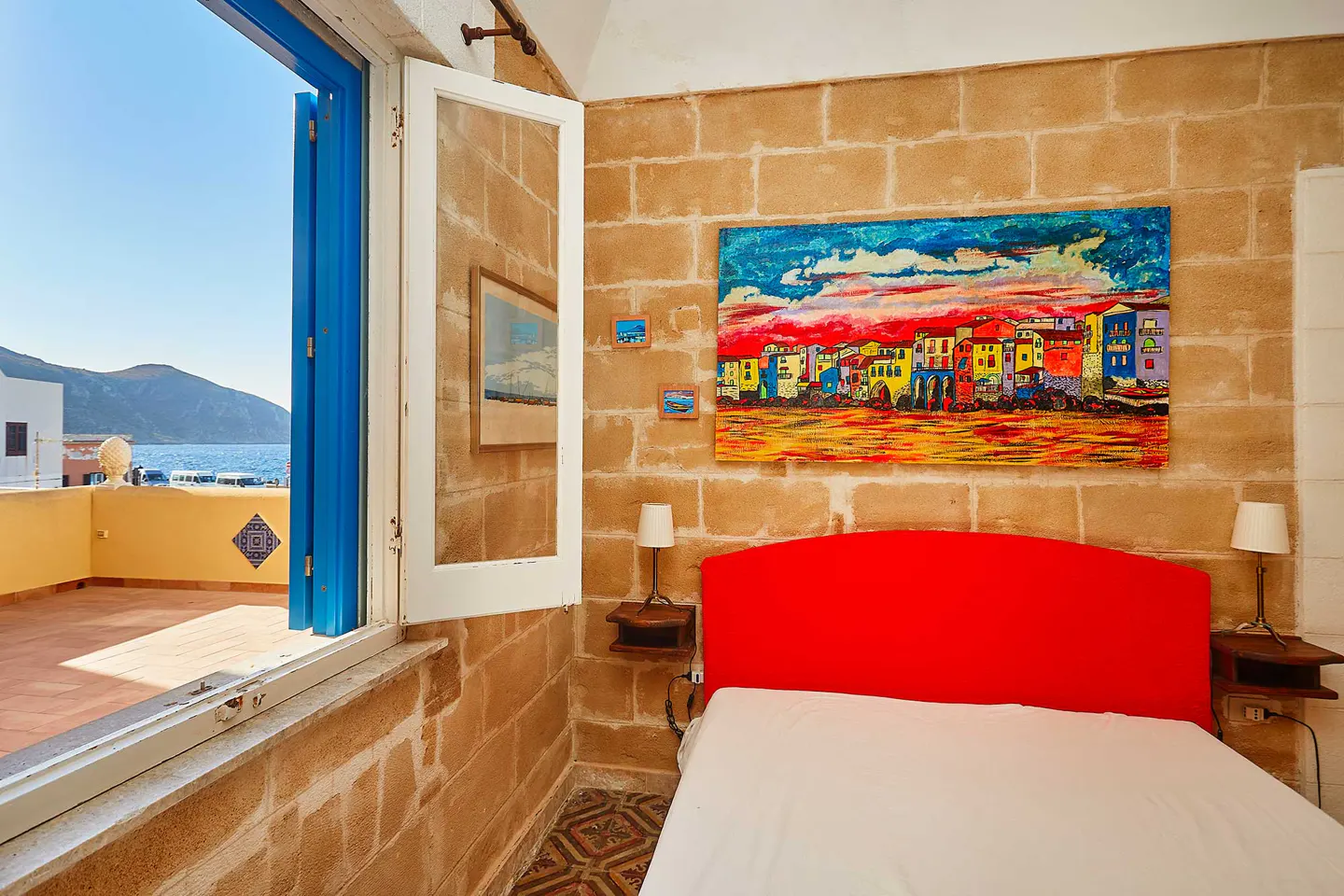 Bedroom with red headboard, stone walls, and open blue door to a balcony overlooking the sea. A colorful painting hangs above the bed.