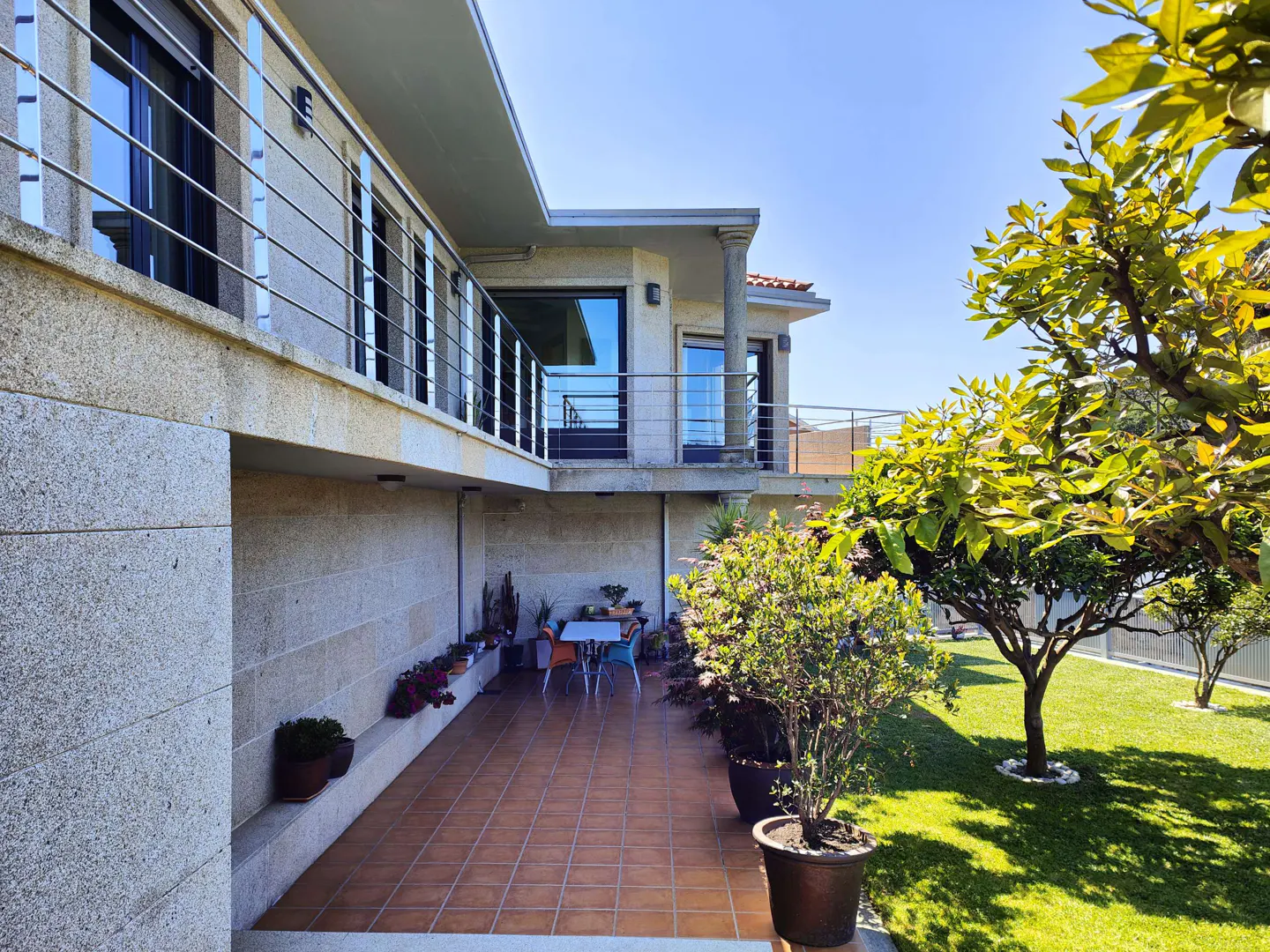 Exterior view of a two-story stone house with a patio, table, and green lawn under a blue sky.
