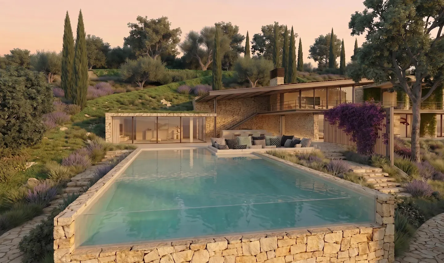 Exterior view of a modern stone house with a pool. The pool is surrounded by stone and lavender. The house has large windows and a patio with seating.