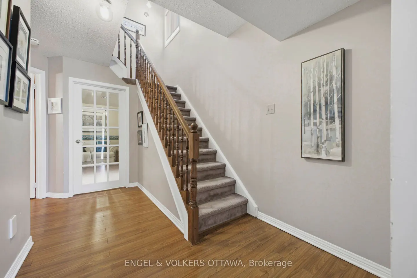 A foyer with hardwood floors, a staircase with brown carpet, and a white door with glass panes. A framed print hangs on the wall.