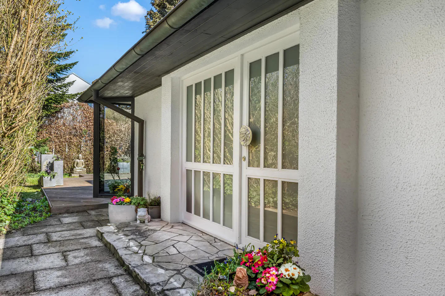 Exterior view of a white house with a stone walkway leading to a glass-paneled door and a covered porch. Flowers are planted near the entrance.