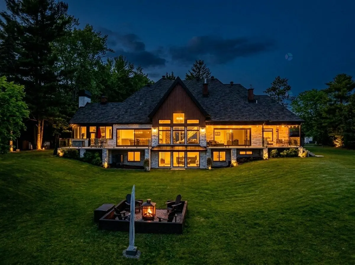 Night view of a large, illuminated stone house with a dark roof, surrounded by green lawn and trees. A fire pit with chairs is in the foreground.