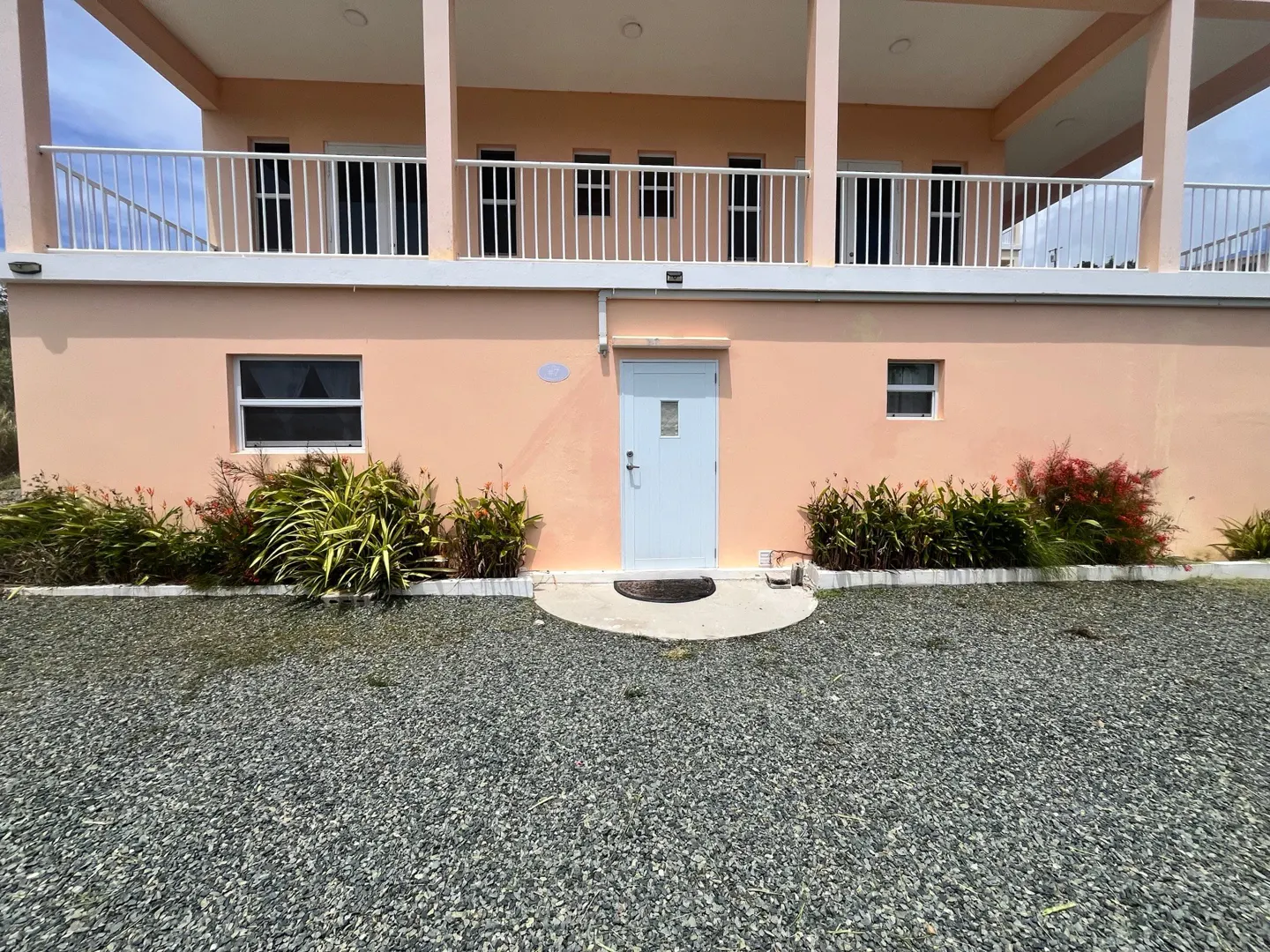Two-story peach building with a white door and small windows. A gravel yard and green plants surround the building.