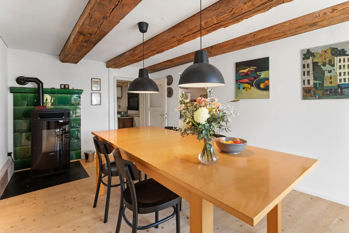 A dining room with a wood table, black chairs, and a vase of flowers. There is a green tiled fireplace and exposed wood beams.