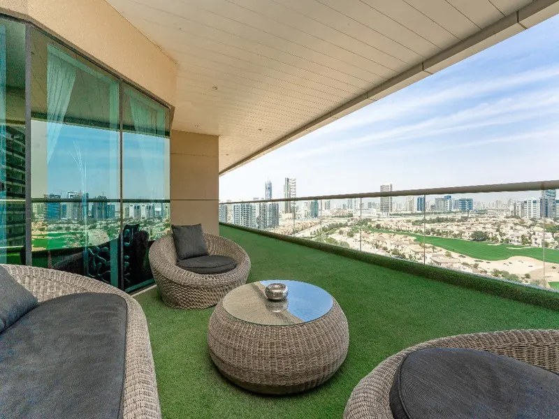 Balcony with wicker furniture, green turf, and city view. Round table and chairs with gray cushions. Glass railing.