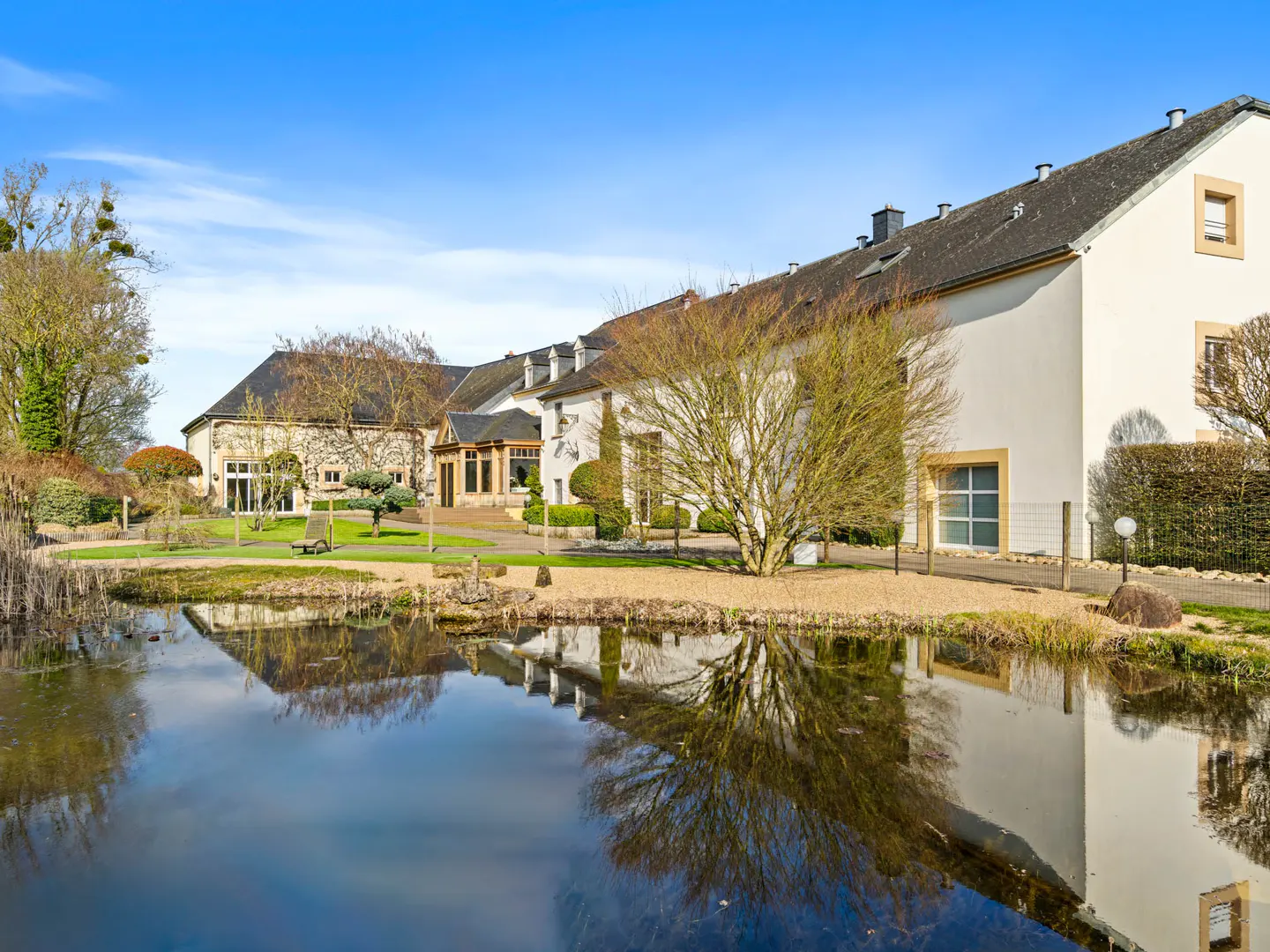 Exterior view of a white building with a dark roof, reflected in a pond under a blue sky. A tree stands in front of the building.