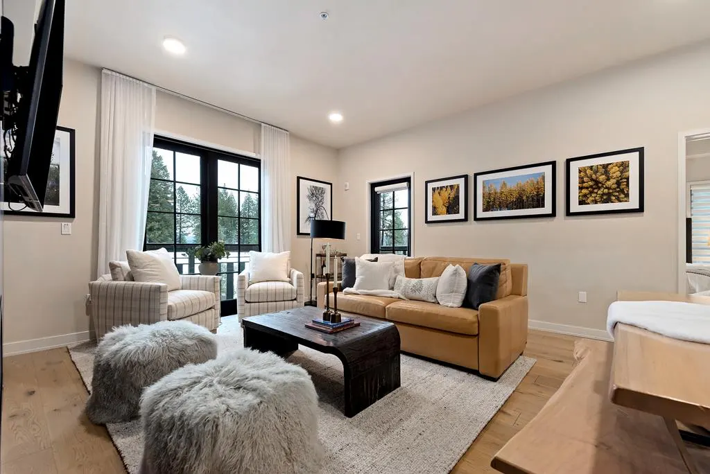 Living room with tan sofa, striped chairs, and fuzzy ottomans on a light rug. Black framed windows and nature art add contrast.