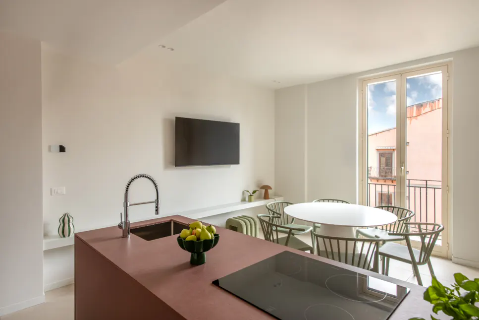 Modern kitchen with a maroon island, stainless steel faucet, and black cooktop. A white table and green chairs sit near a window with a city view.