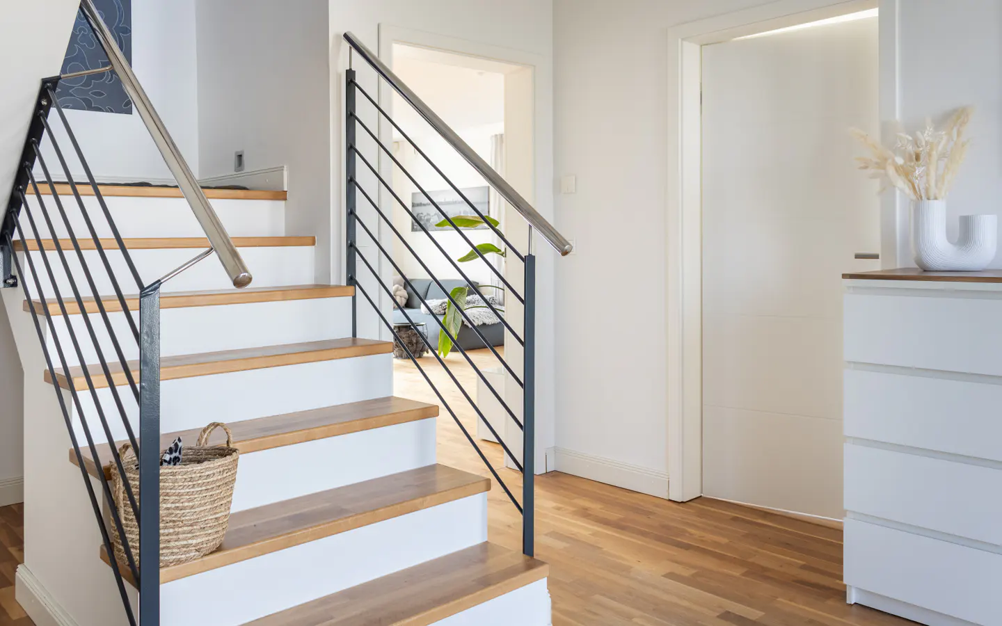 Interior view of a staircase with wood treads and white risers, black metal railing, and a woven basket on the steps. A white dresser and decor are visible.