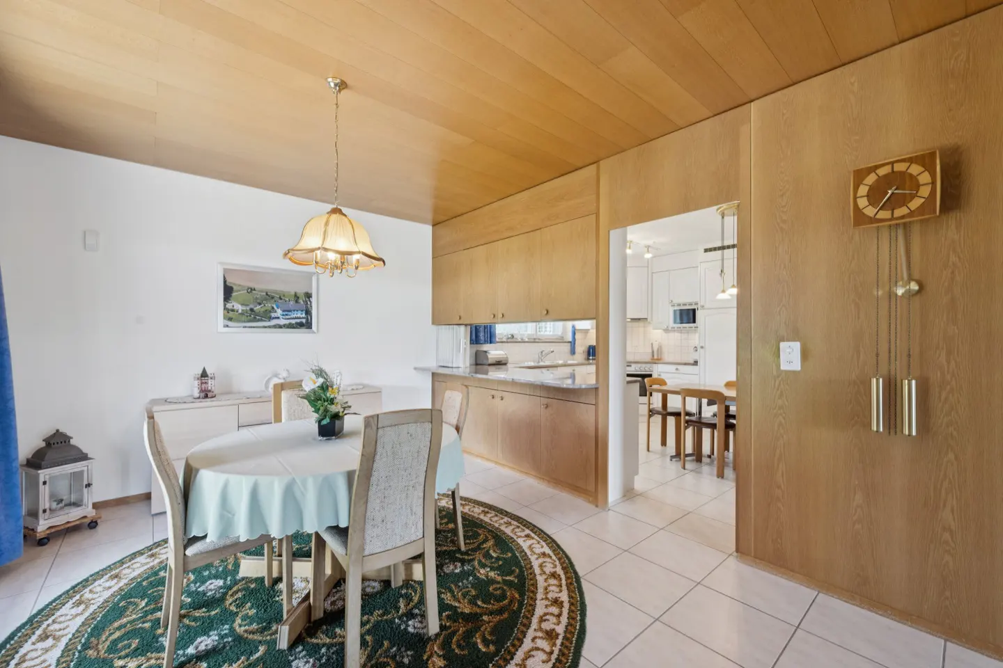 Dining area with round table, chairs, and green rug. Wood paneled walls, ceiling, and a view into the kitchen.