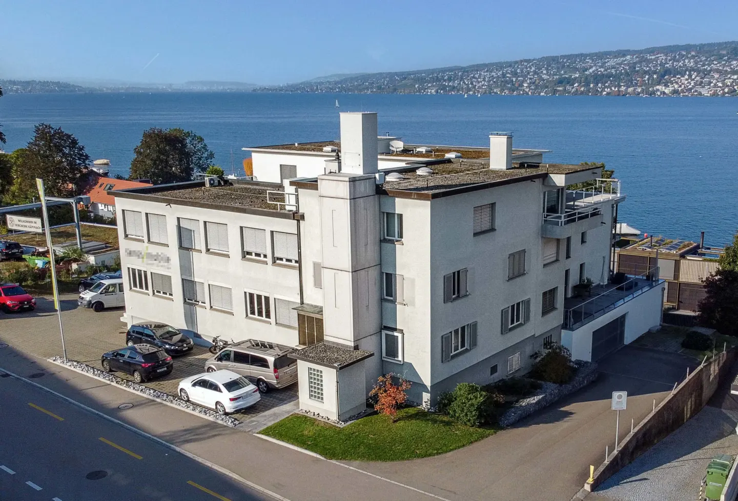 A three-story white building with a flat roof, parked cars, and a lake in the background.
