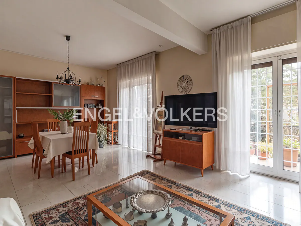 A bright living room with a dining table, TV, and glass doors to a balcony. The room has beige walls, wood furniture, and a patterned rug.