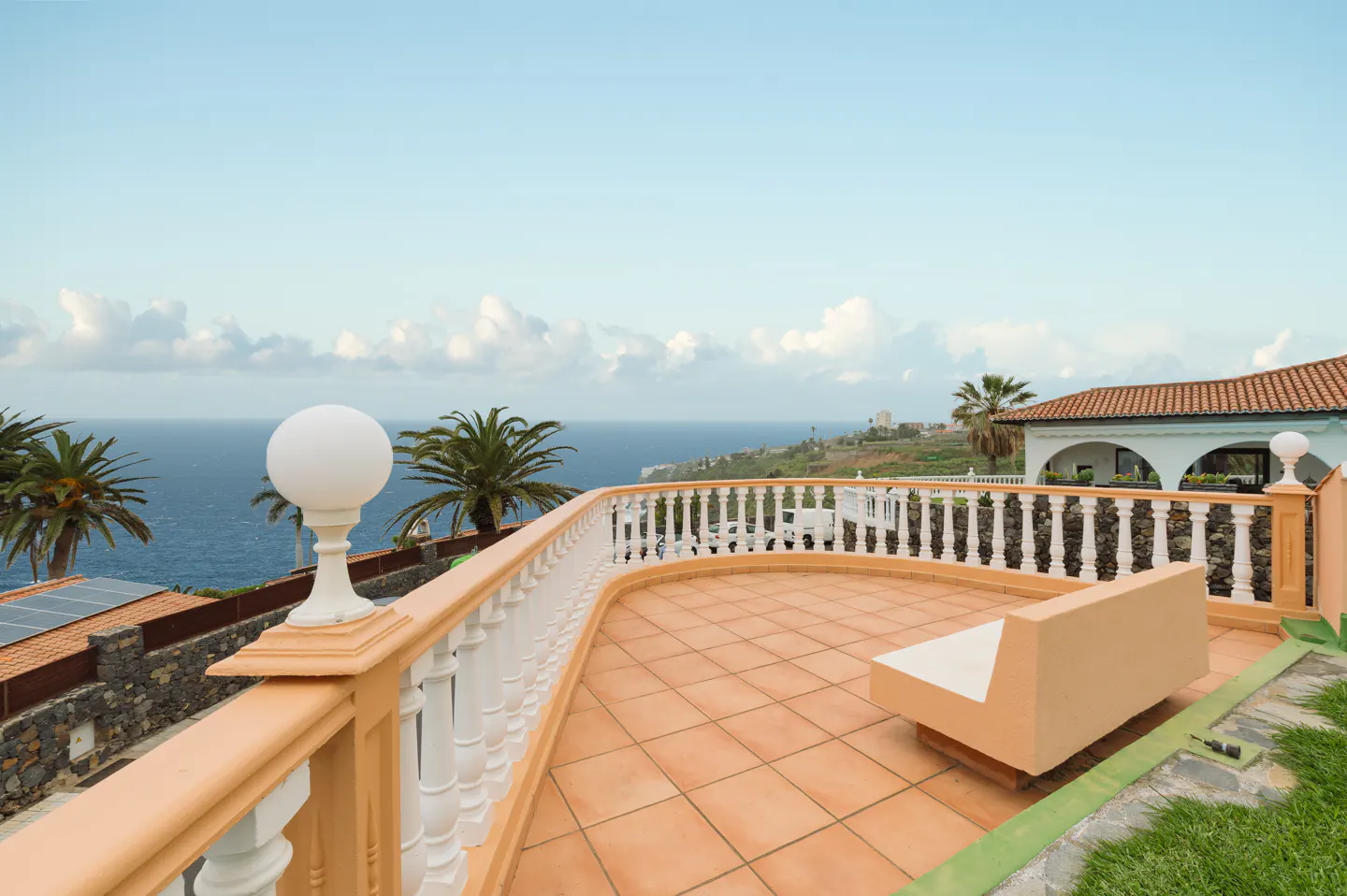 Ocean view from a tiled patio with a white balustrade and peach-colored trim. A white bench sits on the patio, with palm trees and a blue sky in the background.