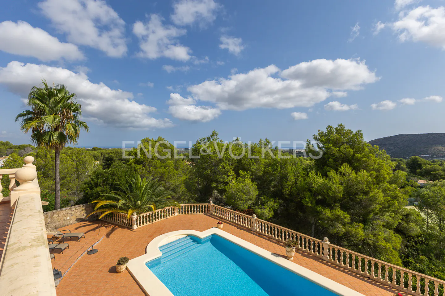 Aerial view of a blue swimming pool with a terracotta deck, surrounded by lush green trees under a blue sky with white clouds.
