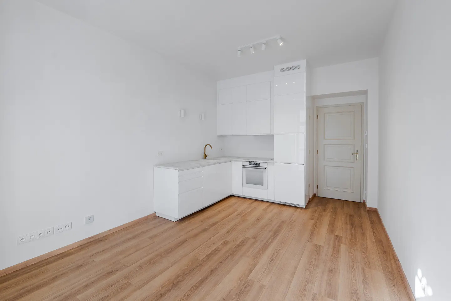 Bright, empty kitchen with white cabinets, marble countertops, and wood floors. A gold faucet and white door add detail.