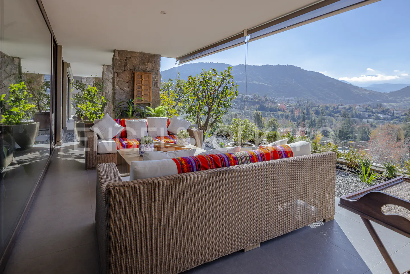 Outdoor patio with wicker furniture, white cushions, and colorful blankets. A mountain view is visible through a glass barrier.