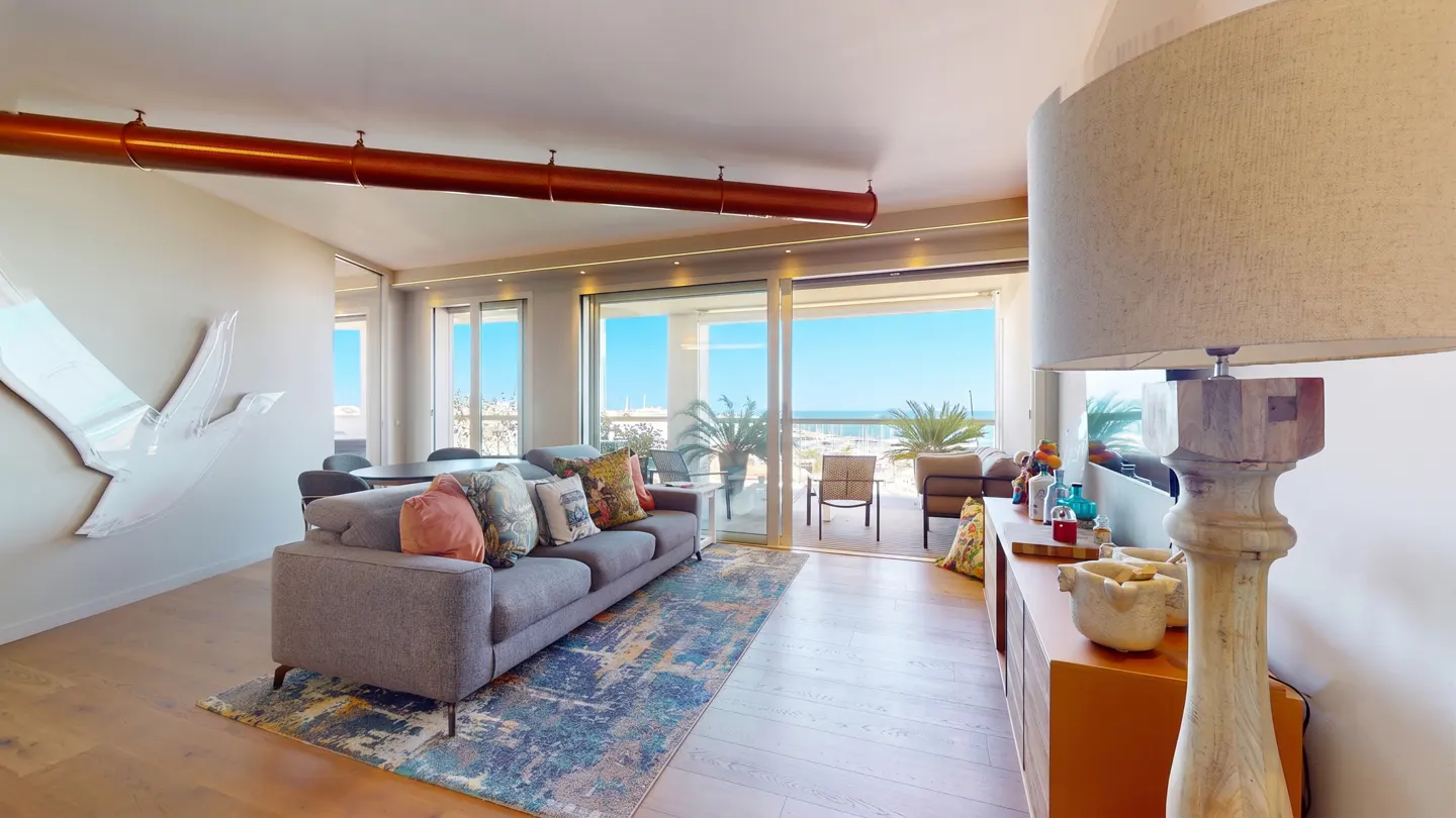 Modern living room with a grey sofa, colorful rug, and large sliding doors opening to a balcony with a sea view.
