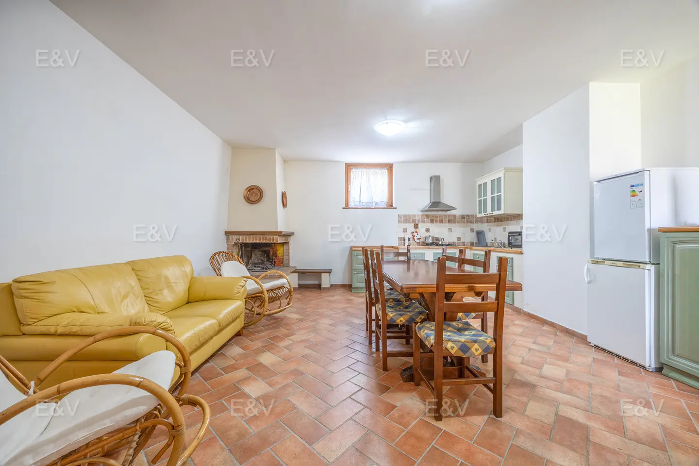 Living room with yellow sofa, fireplace, dining table, and kitchen in the background. The floor is tiled in terracotta color.