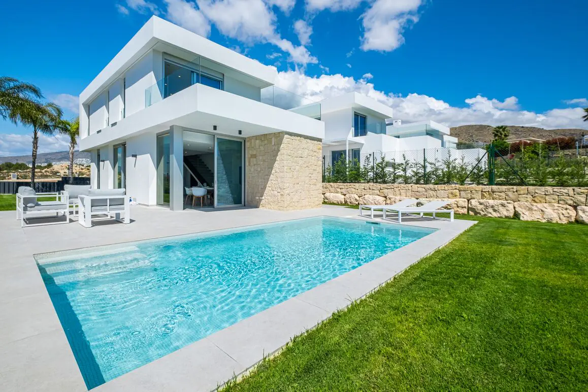 Modern white house with a pool, patio furniture, and green lawn under a blue sky with clouds.