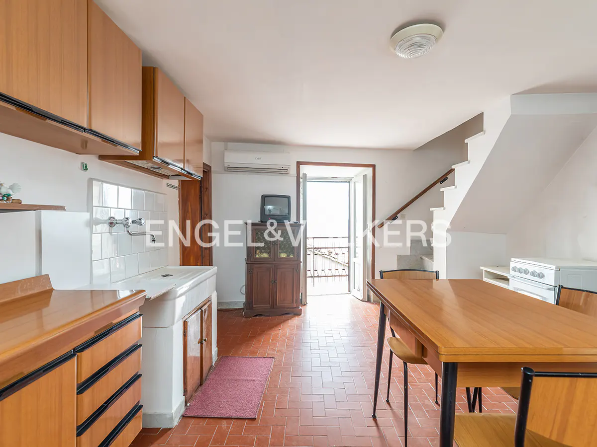 A kitchen and dining area with wood cabinets, a white sink, and a wooden table with chairs. Stairs lead to the upper floor. A balcony door lets in natural light.