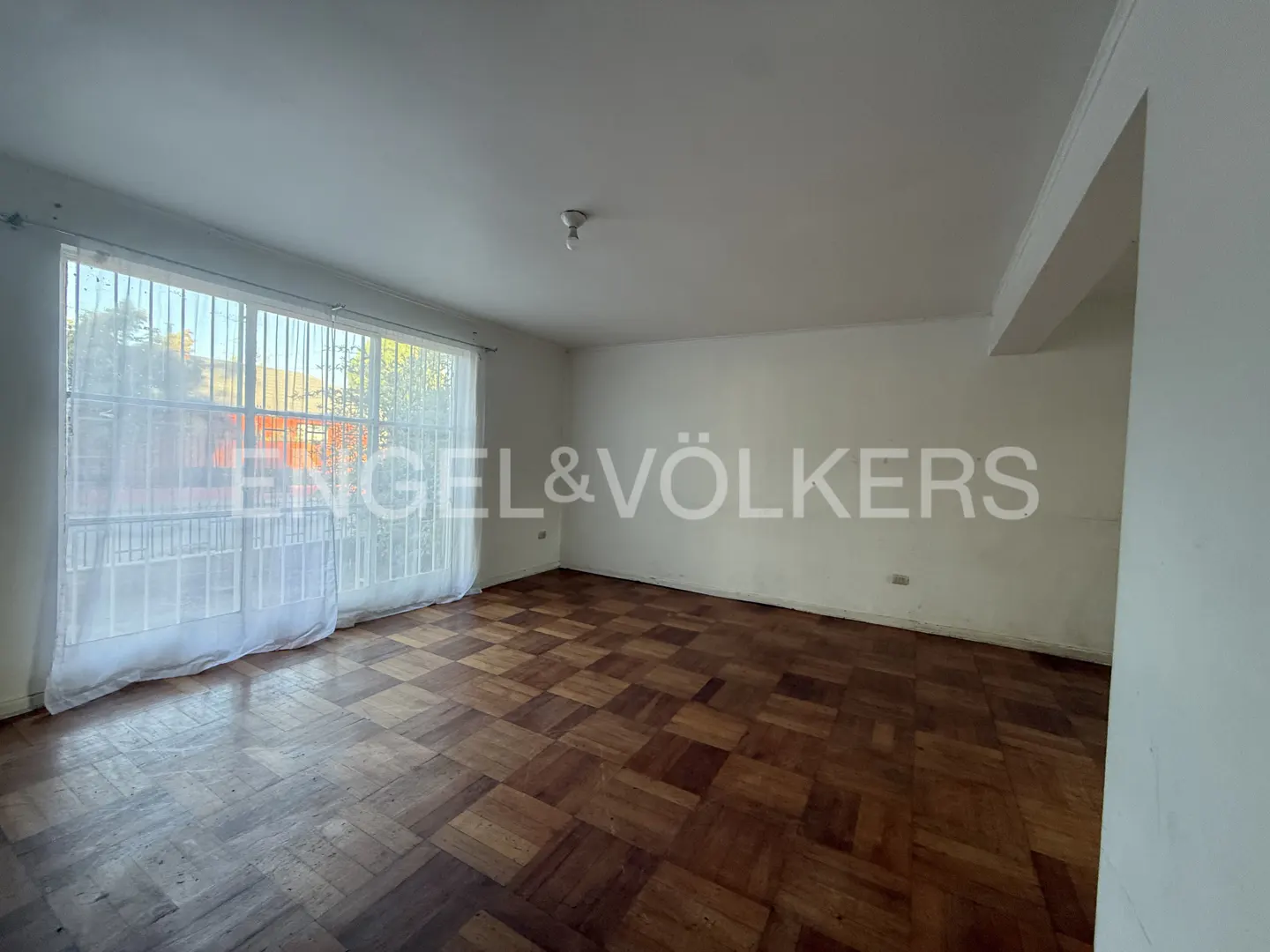 Empty room with parquet floor, white walls, and large window with sheer curtains. Engel & Völkers logo is superimposed on the image.