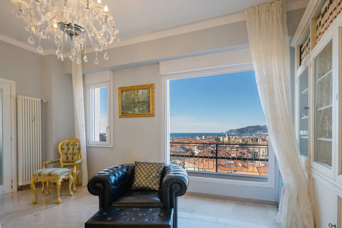 A bright living room with a black leather chair, a gold chair, and a large window with a city view. A chandelier hangs from the ceiling.