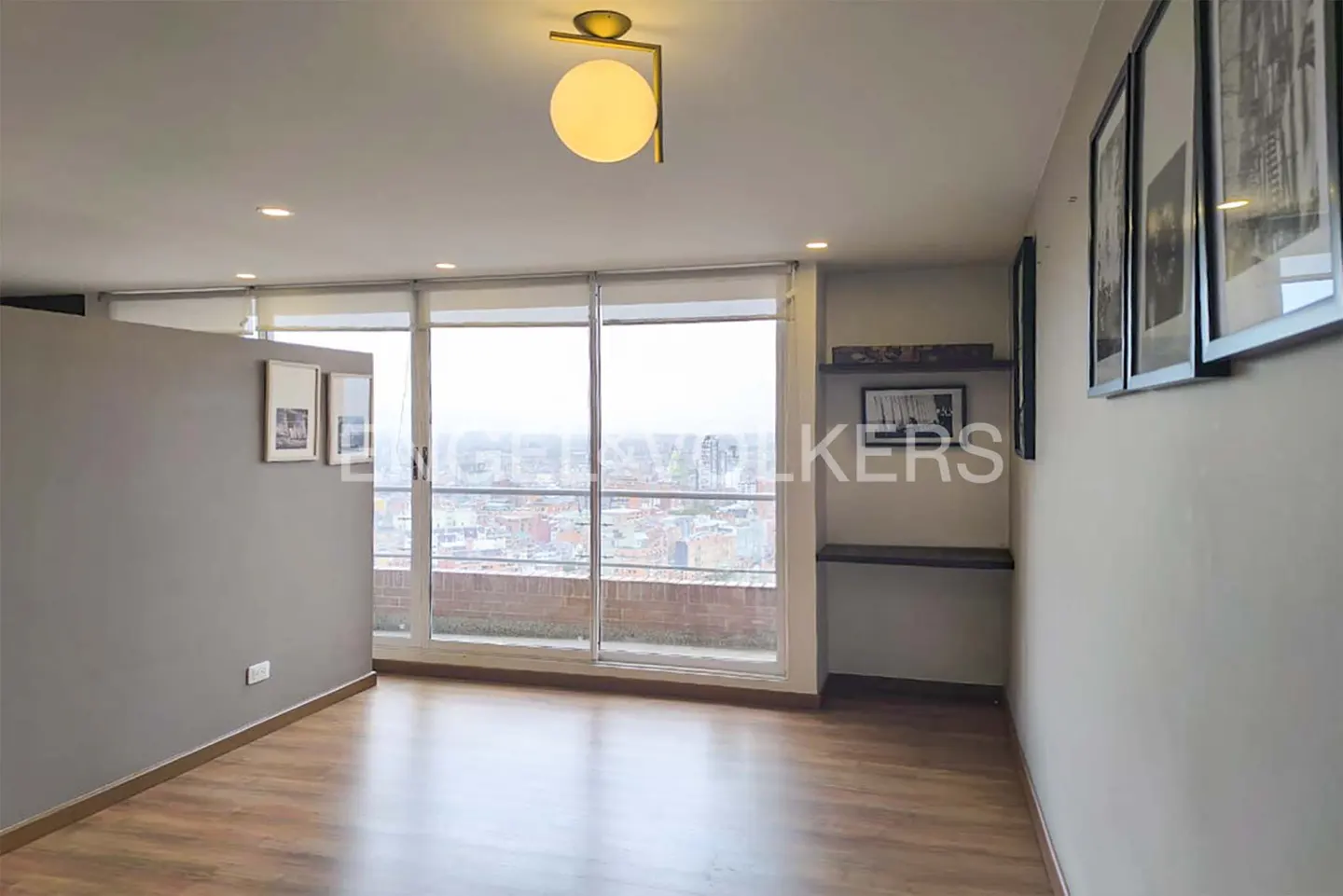 Bright apartment interior with wood floors, gray walls, and large windows overlooking a city. A modern gold light fixture hangs from the ceiling.