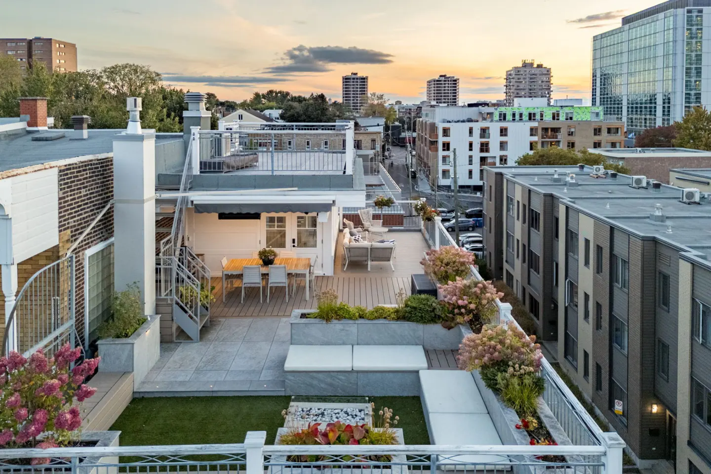 Rooftop terrace with dining area, lounge seating, and planters overlooking a city skyline at sunset.