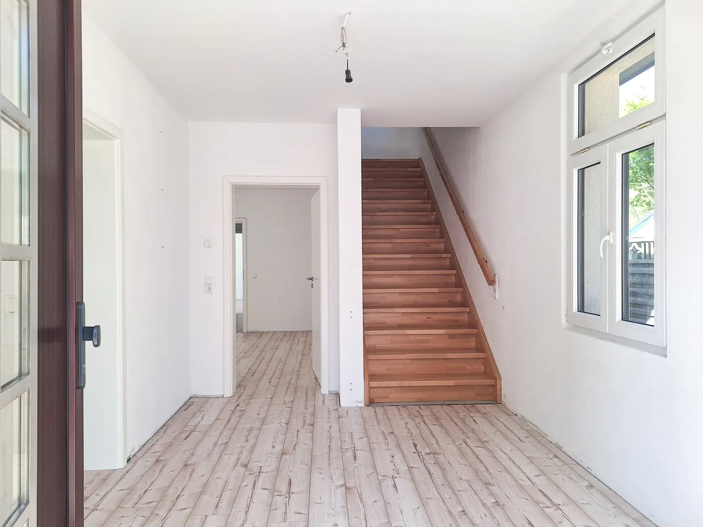 Interior view of a home's entryway with white walls, wood floors, and a staircase. A window and open door are visible.