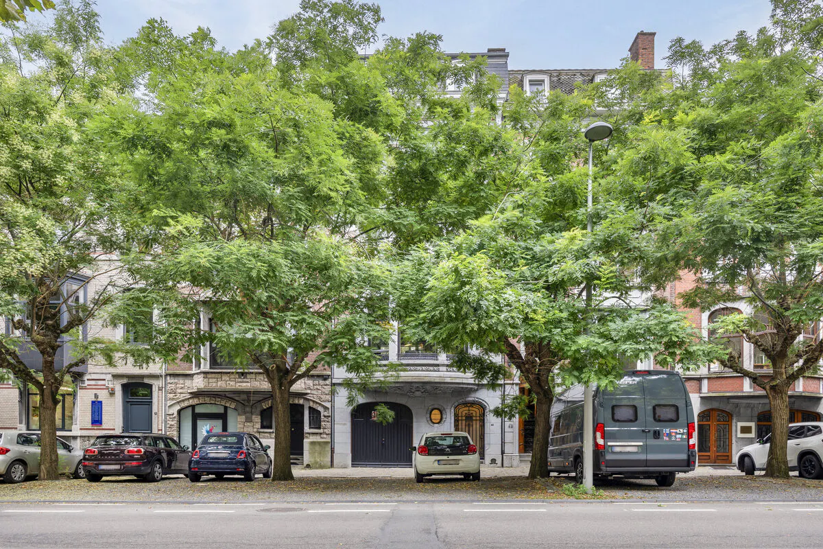 Street view of European-style buildings with cars parked along the curb, framed by green trees.