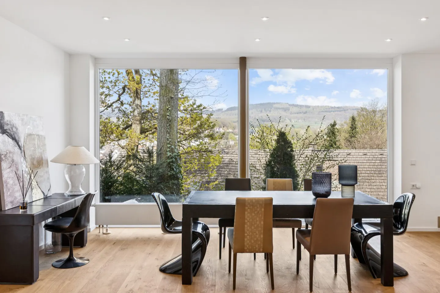 Bright dining room with wood floors, a black table and chairs, and a large window with a view of trees and hills.