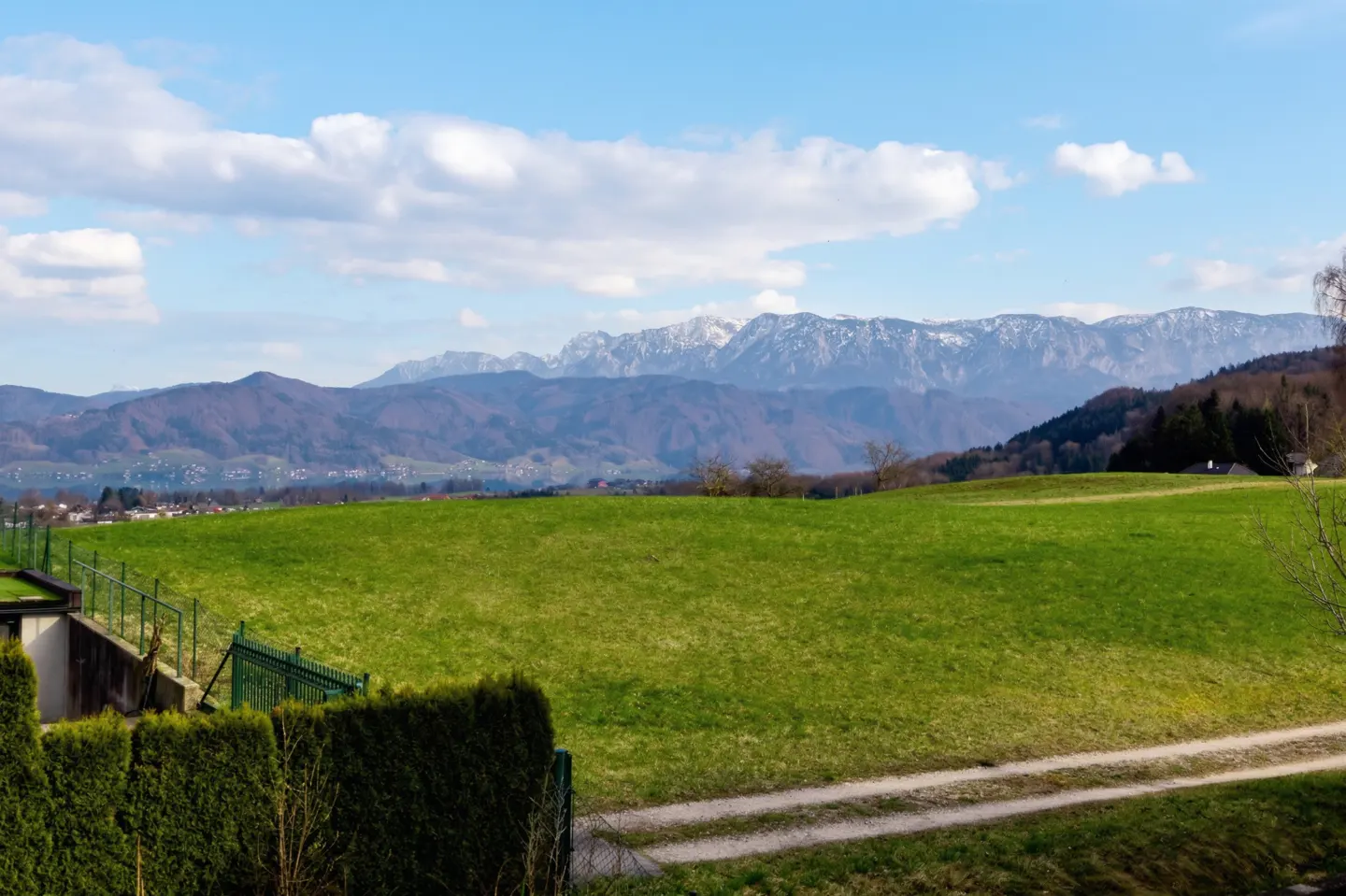 Scenic view of a green field leading to mountains under a blue, cloudy sky. A path cuts through the grass, with trees and a fence in the foreground.