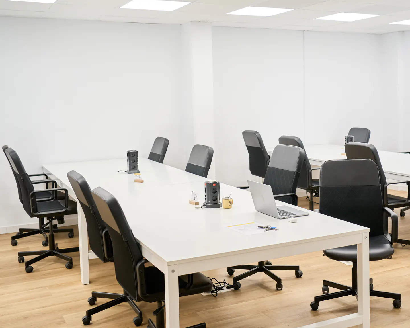 Bright office meeting room with white tables, black ergonomic chairs, and light wood flooring under recessed lighting.