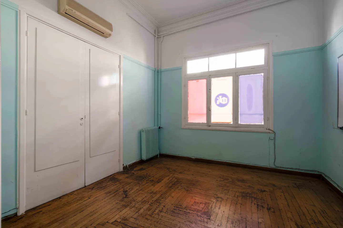 Empty room with parquet floor, light blue walls, and a window with pink, white, and purple stained glass. White double doors and an air conditioner are on the left.