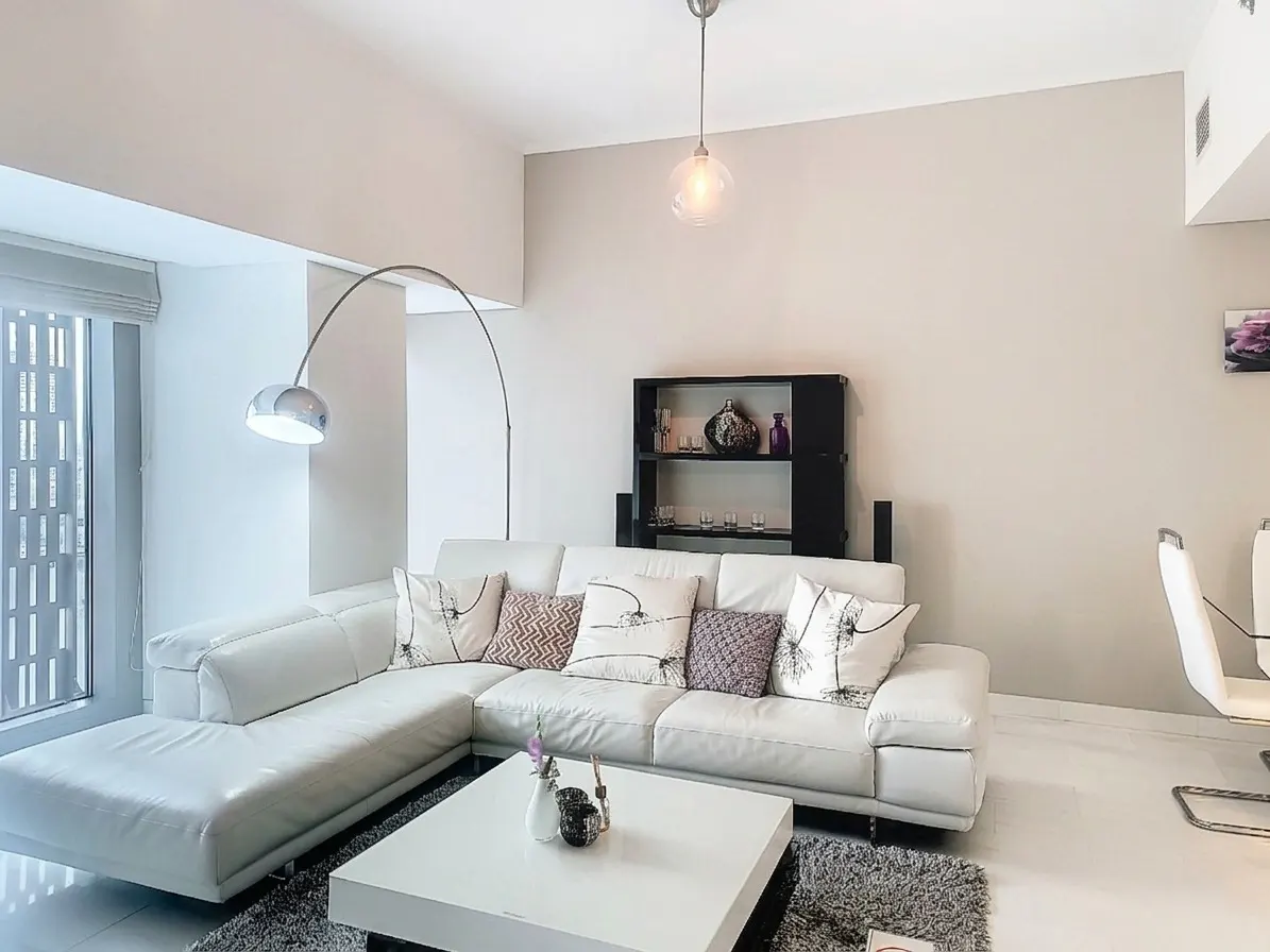 Bright living room with a white sectional sofa, pillows, a white coffee table, and a gray rug. A chrome floor lamp and a black bookshelf are also visible.