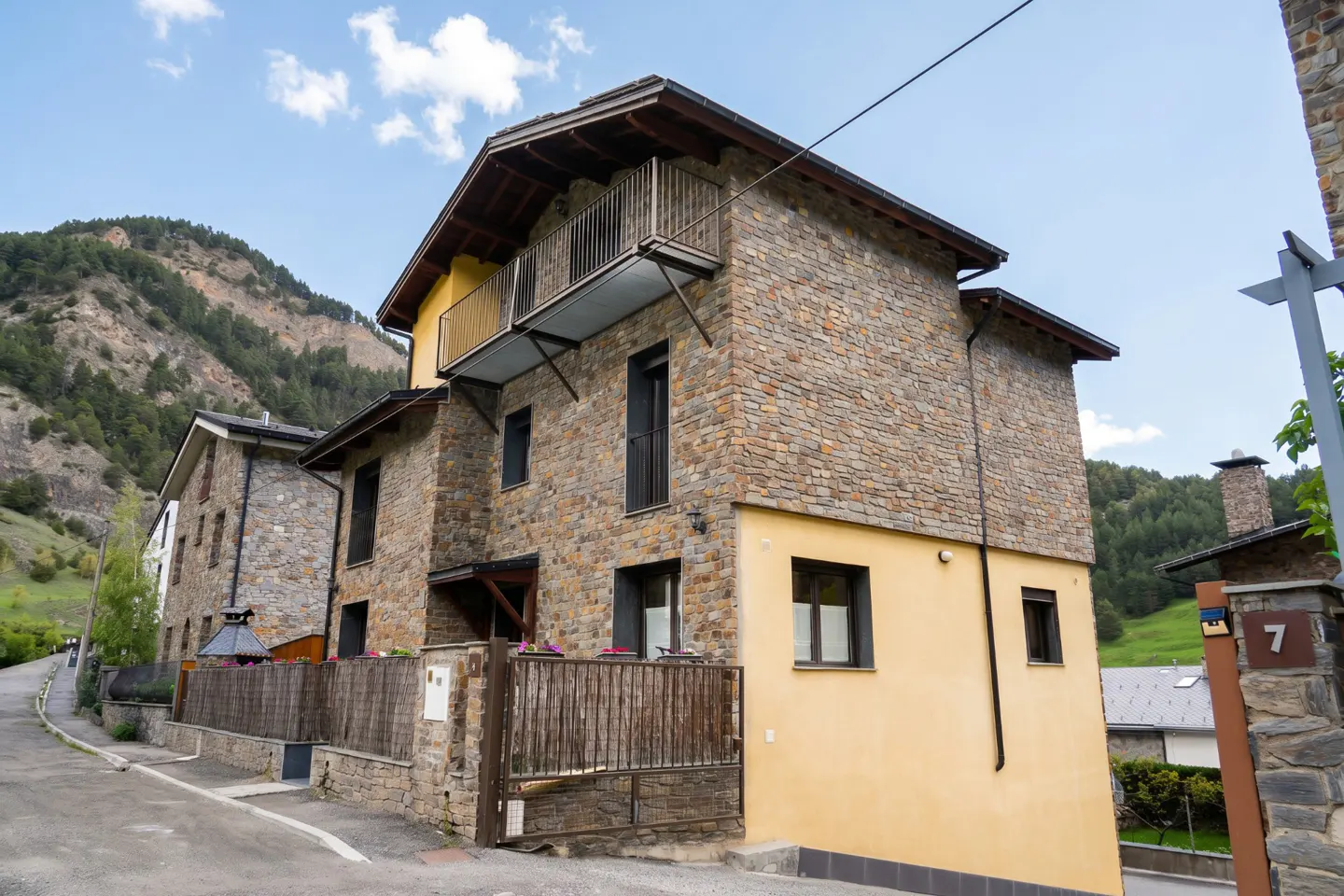 Two-story house with stone and yellow siding, a balcony, and a wooden fence, set against a mountain backdrop.