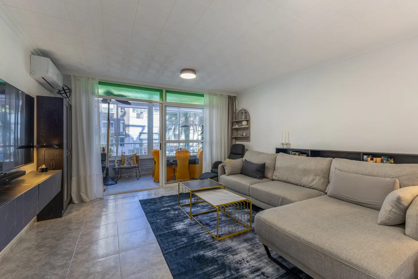 Living room with a beige sectional sofa, a blue rug, and a balcony with yellow chairs.