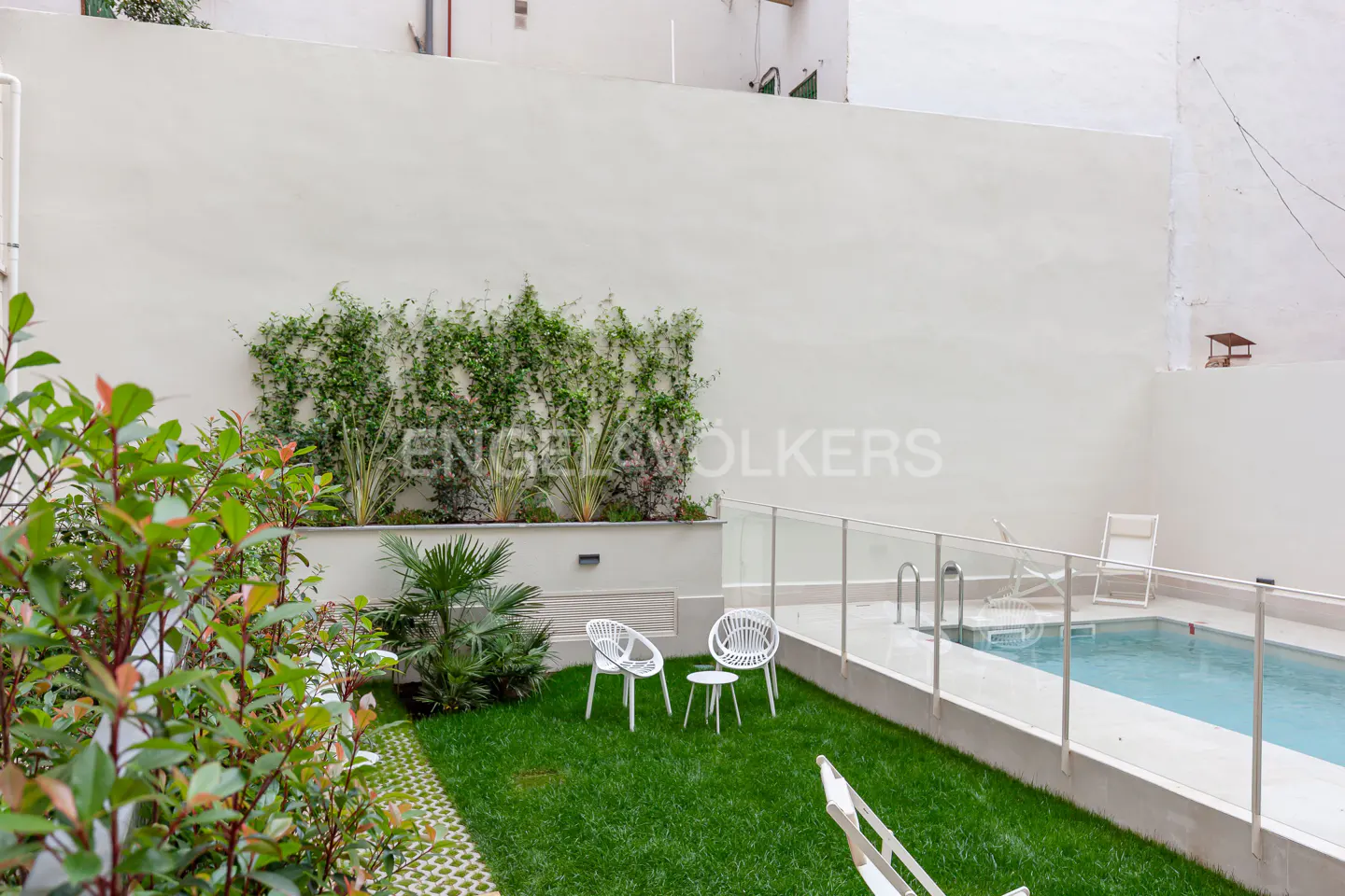 A backyard with a pool, green grass, and white chairs. A white wall is in the background.