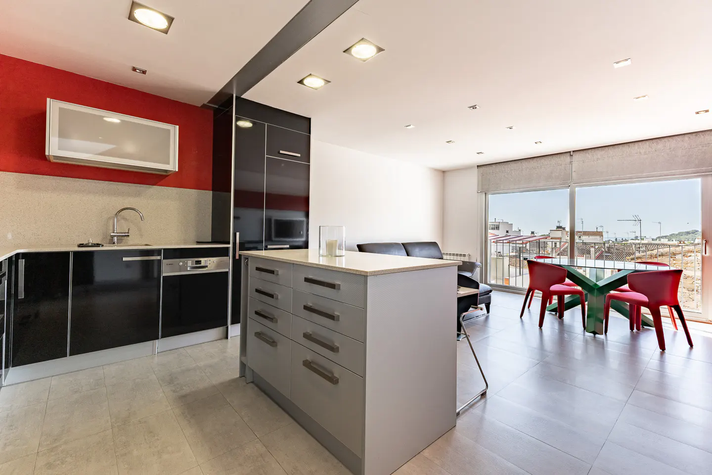 A modern apartment interior featuring a kitchen with black cabinets, a gray island, and a dining area with red chairs. A large window offers a city view.