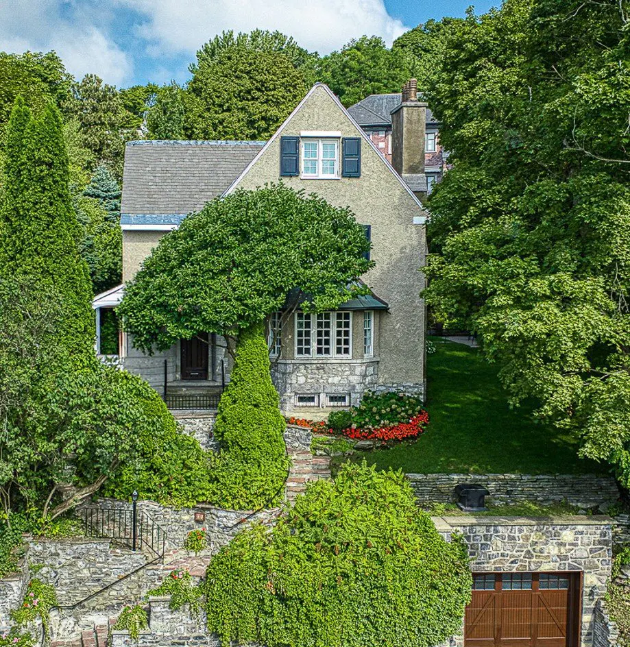A two-story house with a gray roof and stucco walls, surrounded by lush greenery and a stone wall.