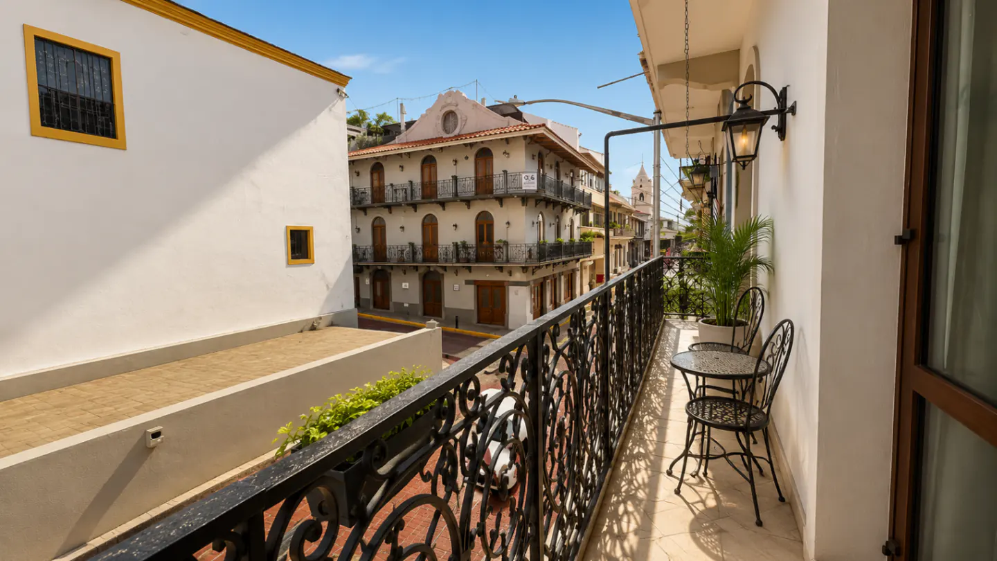View from a balcony with black iron railings, a table and chairs, overlooking a street with old buildings.