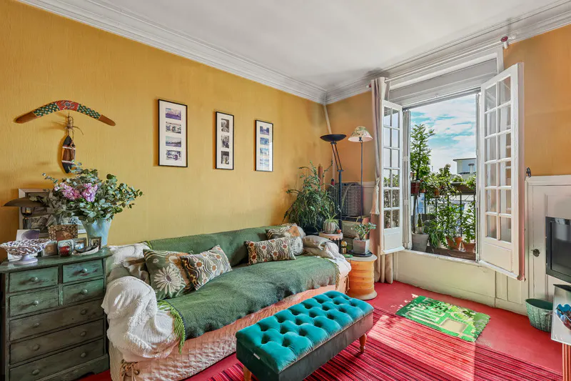 Cozy living room with yellow walls, green sofa, and turquoise ottoman. Balcony with plants visible through open French doors.